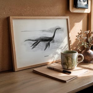 Pode incluir: Uma ilustra&ccedil;&atilde;o em preto e branco emoldurada do Monstro do Lago Ness, rotulada "Nessie", repousa sobre uma superf&iacute;cie de madeira. Uma caneca de cer&acirc;mica com esmalte verde e branco, uma caneta e um bloco de notas tamb&eacute;m est&atilde;o na superf&iacute;cie.