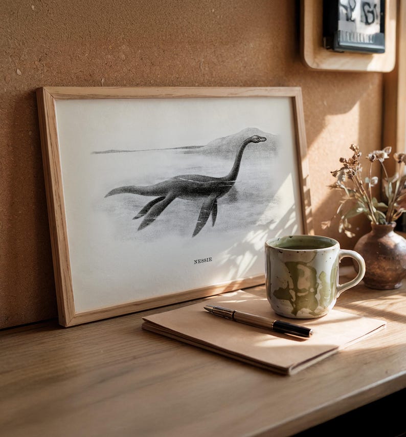 Pode incluir: Uma ilustra&ccedil;&atilde;o em preto e branco emoldurada do Monstro do Lago Ness, rotulada "Nessie", repousa sobre uma superf&iacute;cie de madeira. Uma caneca de cer&acirc;mica com esmalte verde e branco, uma caneta e um bloco de notas tamb&eacute;m est&atilde;o na superf&iacute;cie.