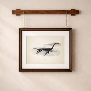 Pode incluir: Ilustra&ccedil;&atilde;o em preto e branco emoldurada do Monstro do Lago Ness, ou Nessie, nadando em um lago. A obra de arte est&aacute; em uma moldura de madeira marrom escura, pendurada em uma barra de madeira com correntes douradas.