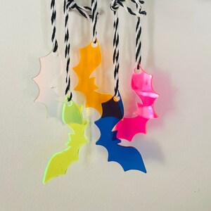 May include: A collection of colorful acrylic bat ornaments in pink, yellow, orange, blue, and green, suspended from black and white twine. The bats are hanging from a white skeleton hand, creating a spooky Halloween theme.