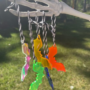 May include: A collection of colorful acrylic bat ornaments hanging from a white skeleton hand. The bats are in various colors, including iridescent, green, yellow, orange, and blue. They are suspended by black and white twine.