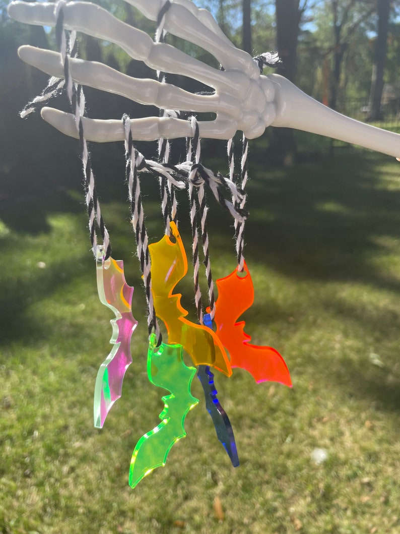 May include: A collection of colorful acrylic bat ornaments hanging from a white skeleton hand. The bats are in various colors, including iridescent, green, yellow, orange, and blue. They are suspended by black and white twine.