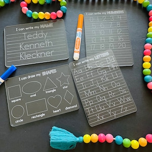 May include: Four clear plastic dry-erase boards with white writing. The boards are for learning letters, numbers, shapes, and writing a name. The boards are on a black surface with a colorful beaded garland.