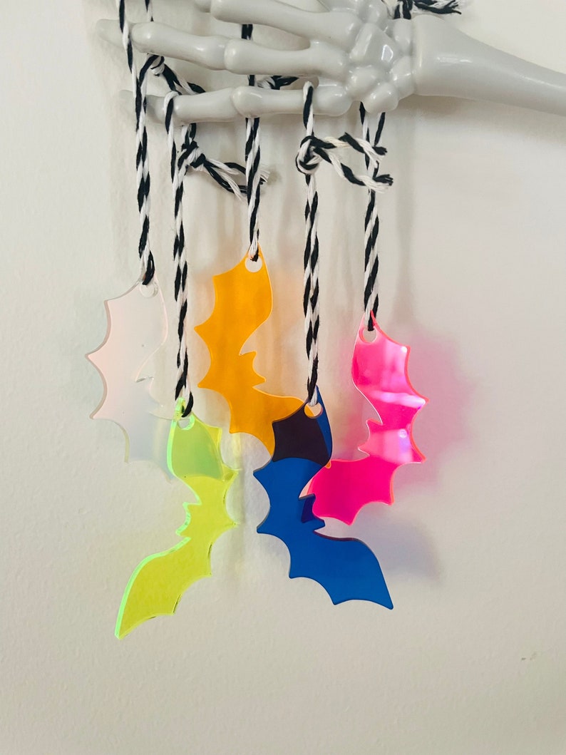 May include: A collection of colorful acrylic bat ornaments in pink, yellow, orange, blue, and green, suspended from black and white twine. The bats are hanging from a white skeleton hand, creating a spooky Halloween theme.
