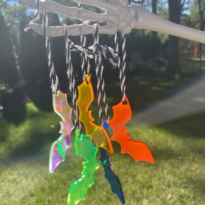 May include: A collection of colorful acrylic bat ornaments suspended from black and white twine. The bats are in shades of orange, green, blue, and iridescent, hanging from a white skeleton hand against a blurred outdoor background.