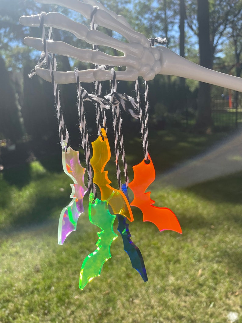 May include: A collection of colorful acrylic bat ornaments suspended from black and white twine. The bats are in shades of orange, green, blue, and iridescent, hanging from a white skeleton hand against a blurred outdoor background.