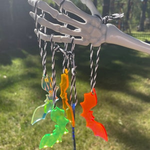 May include: A white skeleton hand holds several colorful acrylic bat ornaments suspended by black and white twine. The bats are various colors, including iridescent, green, orange, and blue. The background is a blurred green lawn and trees.