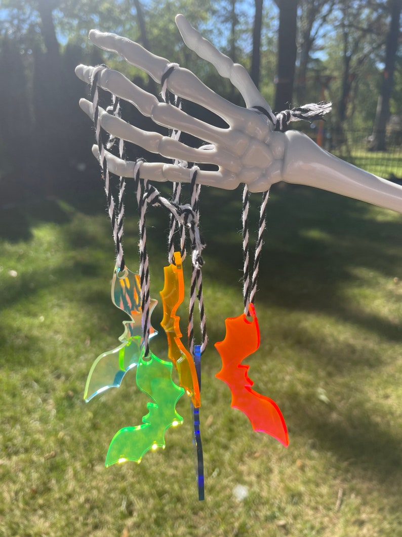 May include: A white skeleton hand holds several colorful acrylic bat ornaments suspended by black and white twine. The bats are various colors, including iridescent, green, orange, and blue. The background is a blurred green lawn and trees.