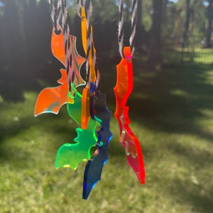 May include: A collection of colorful, translucent bat-shaped ornaments hanging from black and white twine. The bats are in shades of orange, green, blue, and red, with a glossy finish. The background is a blurred view of trees and grass.