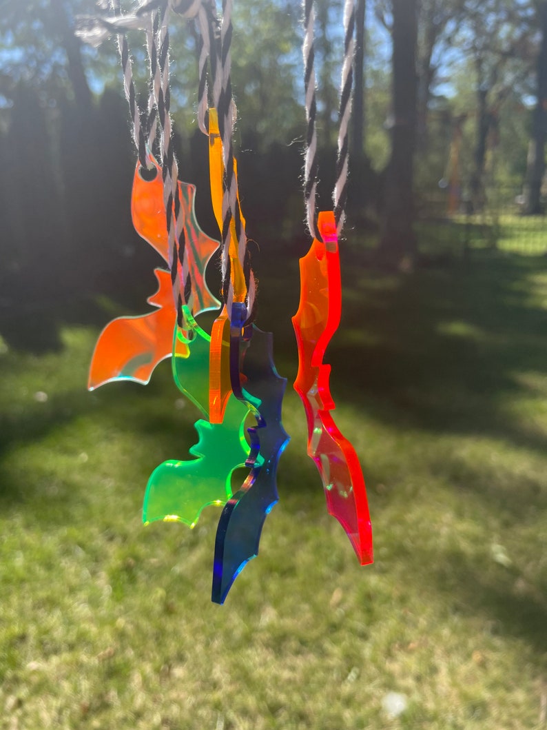 May include: A collection of colorful, translucent bat-shaped ornaments hanging from black and white twine. The bats are in shades of orange, green, blue, and red, with a glossy finish. The background is a blurred view of trees and grass.