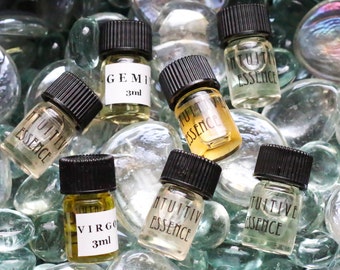 May include: A collection of small glass bottles with black caps, filled with clear and amber liquids. The bottles are labeled with the words "Intuitive Essence", "Gemini 3ml", "Virgo 3ml", and "Jupiter Essence". The bottles are sitting on a bed of clear glass stones.