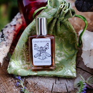 Könnte beinhalten: Ein rechteckiger Glasparfümflakon mit silbernem Verschluss und einem weißen Etikett, das eine schwarze Strichzeichnung einer geflügelten Figur sowie den Text LIQUID PIXIE INTUITIVE ESSENCE zeigt, vor einem hellgrünen Stoffbeutel.