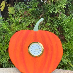 May include: Orange pumpkin-shaped clock with a gold-rimmed clock face and black hands. The clock is made of wood and has a painted orange finish.
