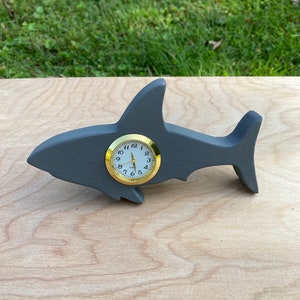 May include: A gray wooden shark clock with a gold-colored face and black hands. The clock is sitting on a light brown wooden surface.