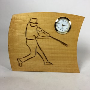 May include: A wooden clock with a carved baseball player design. The clock face is white with black numbers and hands. The clock is mounted on a curved wooden base.