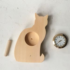 May include: A wooden cat-shaped clock stand with a round hole in the center. A small round clock with gold trim and white face is shown next to the stand.