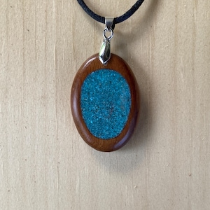 Pode incluir: Um pingente de formato oval com uma moldura de madeira castanha polida que envolve uma incrusta&ccedil;&atilde;o texturizada de um turquesa vibrante. O pingente est&aacute; suspenso por um cord&atilde;o preto simples com um passador de metal prateado, criando um contraste entre a madeira natural e o centro azul-esverdeado.