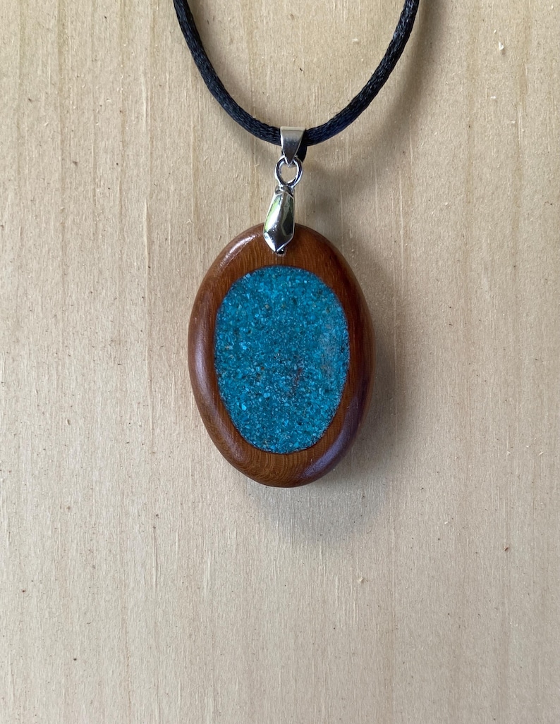Pode incluir: Um pingente de formato oval com uma moldura de madeira castanha polida que envolve uma incrusta&ccedil;&atilde;o texturizada de um turquesa vibrante. O pingente est&aacute; suspenso por um cord&atilde;o preto simples com um passador de metal prateado, criando um contraste entre a madeira natural e o centro azul-esverdeado.