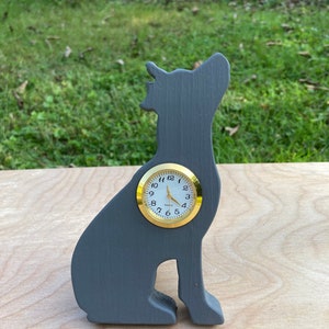 May include: A gray wooden clock shaped like a dog sitting on a wooden surface. The clock face is white with black numbers and gold hands.