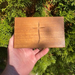 May include: A rectangular wooden box with a carved cross design on the lid. The box is a warm brown color and appears to be made of wood. The box is held in a person's hand against a backdrop of green foliage.
