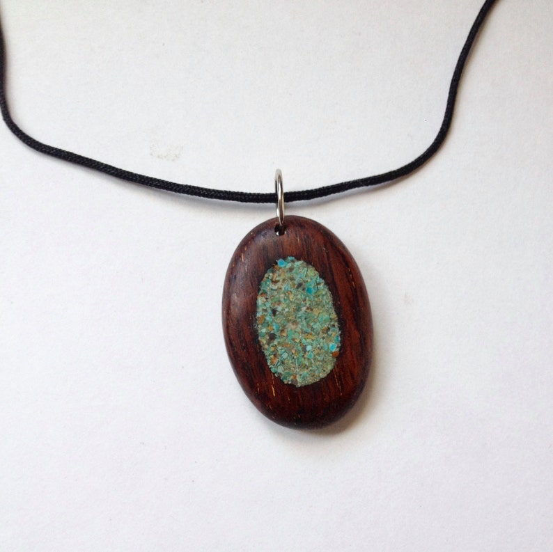 May include: Oval wooden pendant with a turquoise inlay, suspended from a black cord. The pendant has a dark brown wood frame and a central oval of crushed turquoise stones. The necklace is displayed against a white background.