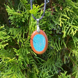 Pode incluir: Um pendente oval com uma moldura de madeira lisa em castanho-avermelhado que rodeia um centro azul-turquesa texturado e cintilante. O pendente est&aacute; preso a um suporte de metal prateado e suspenso por um colar de cord&atilde;o preto fino.