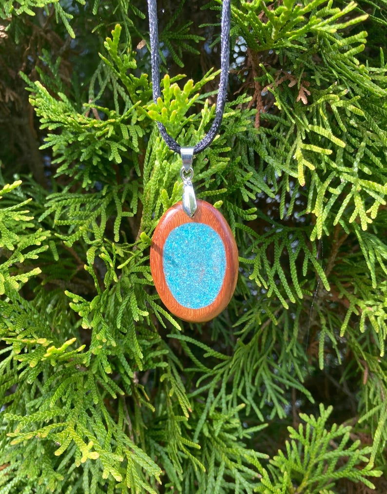 Pode incluir: Um pendente oval com uma moldura de madeira lisa em castanho-avermelhado que rodeia um centro azul-turquesa texturado e cintilante. O pendente est&aacute; preso a um suporte de metal prateado e suspenso por um colar de cord&atilde;o preto fino.