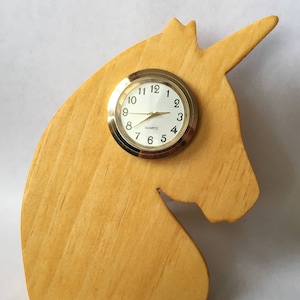 May include: A wooden unicorn-shaped clock with a gold-colored clock face. The clock face has black numbers and hands. The unicorn is made of light-colored wood.