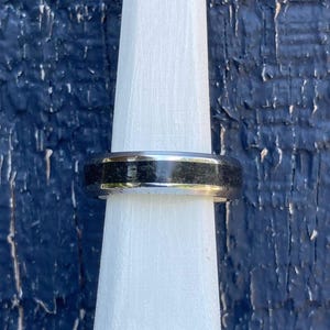 May include: A polished silver-toned metal ring featuring a wide, textured black inlay band that runs horizontally around the center of the ring. The band has a smooth, reflective metallic finish on the top and bottom edges, creating a contrast.