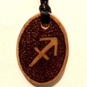 Puede incluir: Colgante ovalado de madera con el símbolo de Sagitario en marrón claro sobre un fondo marrón oscuro texturizado. El colgante cuelga de un cordón negro. Un accesorio con temática zodiacal.