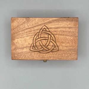 May include: A rectangular wooden box with a hinged lid. The box is made of light brown wood and features a Celtic knot design carved into the top. The box is closed and has a small metal clasp.