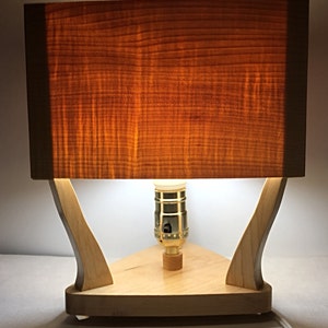 May include: A wooden table lamp with a rectangular lampshade and a triangular base. The lampshade is made of a warm-toned wood, and the base is a lighter wood. The lamp has a standard light bulb socket and a power cord.