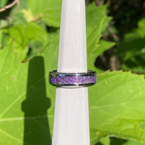May include: A silver-toned metal ring featuring a central inlay of crushed purple material. The band has a smooth, polished finish on the edges, framing the textured, vibrant violet inlay that runs horizontally around the circumference of the ring.