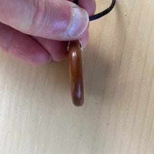 Pode incluir: Um pingente de madeira polida de cor castanho-avermelhada em forma de anel arredondado. O pingente est&aacute; preso a um cord&atilde;o preto fino por uma pequena argola de metal prateado. O veio da madeira &eacute; vis&iacute;vel, apresentando um tom quente.