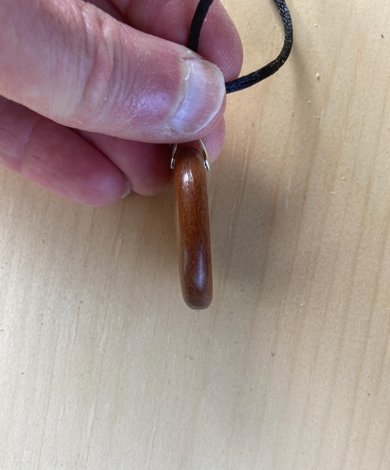 Pode incluir: Um pingente de madeira polida de cor castanho-avermelhada em forma de anel arredondado. O pingente est&aacute; preso a um cord&atilde;o preto fino por uma pequena argola de metal prateado. O veio da madeira &eacute; vis&iacute;vel, apresentando um tom quente.