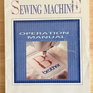 May include: A Brother sewing machine operation manual with a blue and white striped cover. The cover features a photo of a Brother sewing machine embroidering the letter "B" with the word "brother" below it. The manual is titled "Sewing Machine Operation Manual".
