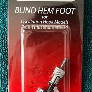 May include: A Janome blind hem foot for oscillating hook models, packaged in a red and black card. The sewing machine accessory is silver and black, designed for creating invisible hems. The packaging includes recycling symbols.