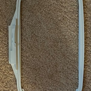 May include: A white plastic embroidery hoop with a 5x7 inch opening. The hoop is designed for use with embroidery machines.