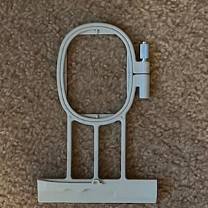 Op de afbeelding: Een witte plastic borduurring met een scharnierende klem. De ring is ontworpen om stof strak te houden voor borduurprojecten.