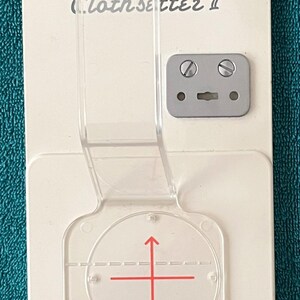 May include: A white Memory Craft Clothsetter II tool with a clear plastic guide. The tool has a gray metal plate with two screws and a clear plastic guide with red crosshairs. The text "Memory Craft Clothsetter II" is printed on the tool.