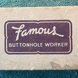 May include: A rectangular box with the words "Famous BUTTONHOLE WORKER" in a dark purple, framed by a white border. The box has a light blue-gray background with some discoloration.