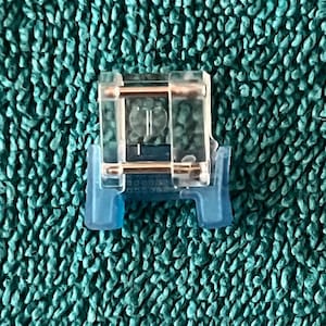 May include: A clear plastic sewing machine foot with a metal bar and blue plastic feet. The foot is designed to attach to a sewing machine and guide fabric during stitching. The image is set against a textured teal background.