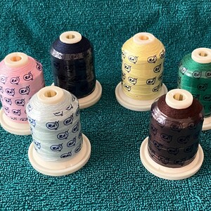 May include: Six spools of colorful embroidery thread are arranged on a teal surface. The spools are in various colors, including pink, yellow, green, brown, blue, and light blue. Each spool has a white base and top, and is decorated with a repeating pattern.