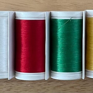 May include: Seven spools of thread in different colors: black, white, red, green, yellow, and blue. The spools are arranged in a row on a wooden surface.