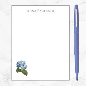 May include: A white notepad with the name "ANNA FAULKNER" printed at the top. A blue pen is on the right side of the notepad. A blue hydrangea flower is in the bottom left corner of the notepad.