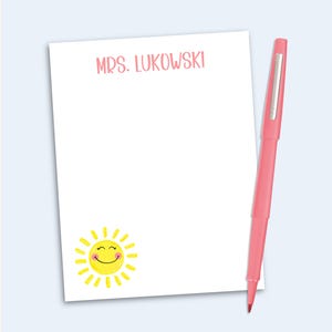 May include: A white rectangular notepad featuring the text MRS. LUKOWSKI in pink at the top and a yellow smiling sun graphic in the bottom left corner, accompanied by a pink ballpoint pen with a silver clip positioned vertically on the right side.