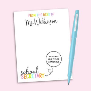 May include: A white notepad with the text FROM THE DESK OF Ms. Wilkenson in colorful letters and school secretary in black and colorful script. A light blue pen is positioned to the right of the notepad. A black circle with the text MULTIPLE JOB TITLES AVAILABLE points to the text with an arrow.