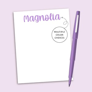 May include: A white notepad with the word Magnolia printed in purple script at the top. A circular graphic with an arrow points to the text, containing the words MULTIPLE COLOR CHOICES. A purple pen with a silver clip is positioned to the right.
