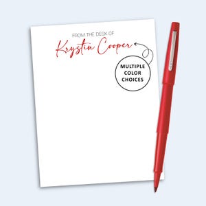 May include: A white rectangular notepad with the text FROM THE DESK OF Krystin Cooper in red and black ink, accompanied by a red ballpoint pen. A black circular graphic with an arrow points to the name, containing the text MULTIPLE COLOR CHOICES.
