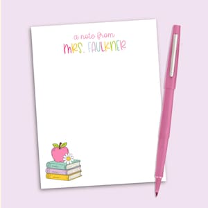May include: A white notepad with the text "a note from MRS. FAULKNER" in colorful letters at the top and a graphic of three stacked books topped with a pink apple and a white daisy in the bottom left corner, accompanied by a pink pen.